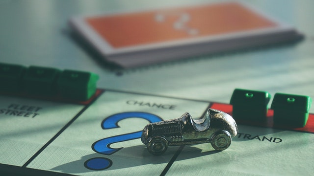 Foto do jogo Monopoly contendo um pedaço do tabuleiro, 5 casas em dois espaços diferentes, um carro de plástico e um baralho de cartas