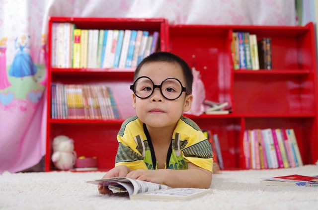 menino oriental com óculos de armação redondos, lendo uma revista, olhando para a câmera. Fundo com estante livreiro vermelha, com livros infantis